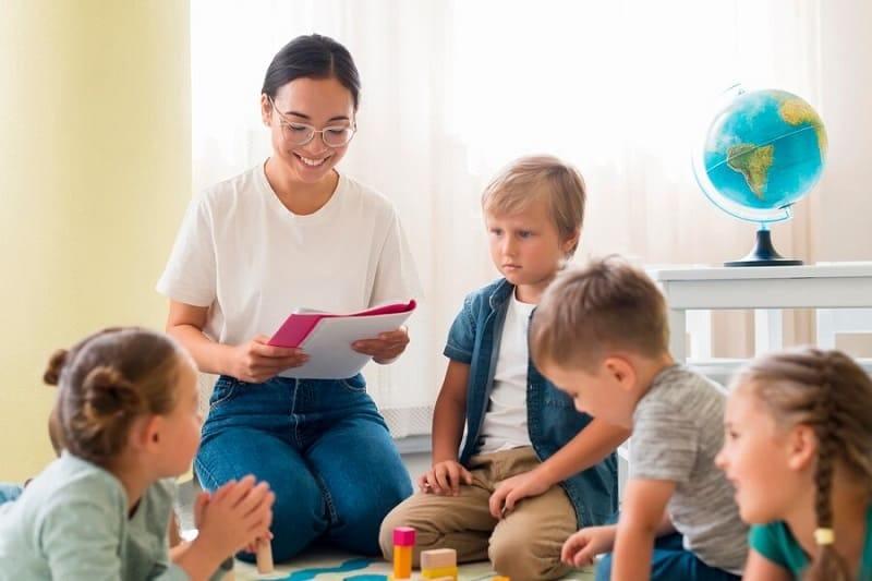Một giáo viên mỉm cười nói chuyện với một nhóm học sinh tiểu học đang chăm chú lắng nghe trong lớp học đầy màu sắc, thể hiện sự kết nối và tương tác tích cực.